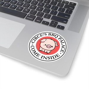 May include: A round sticker with the text "CIRCE'S BBQ PALACE COME INSIDE" encircling a cartoon pig on a red background. The sticker is white with black text and is placed on a laptop.