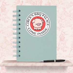 May include: A round sticker with the text "CIRCE'S BBQ PALACE COME INSIDE" encircling a cartoon pig illustration. The pig is pink with a black snout and is centered on a red circle. The sticker is on a light blue notebook.