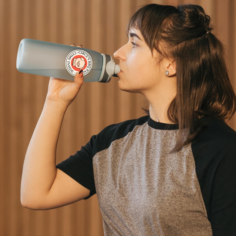May include: A light blue water bottle with a gray cap and a circular sticker that reads "Inside Come's BBQ Pal". The person is drinking from the bottle. The person is wearing a gray and black raglan shirt.