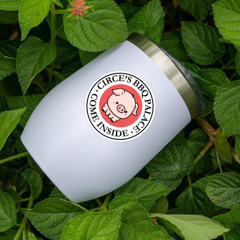 May include: A white insulated tumbler with a silver rim, featuring a circular sticker. The sticker has a red background with the text "Circe's BBQ Palace" and "Come Inside" surrounding a cartoon pig.