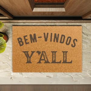 Puede incluir: Una alfombra rectangular con las palabras "BEM-VINDOS Y'ALL" en letras marrones oscuras. La alfombra es de color marrón claro y está colocada frente a una puerta de madera.