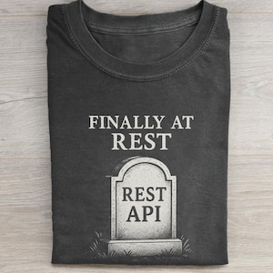 May include: Dark gray t-shirt with the text "FINALLY AT REST" above a tombstone that reads "REST API". The tombstone is a light gray color and has a simple design. The shirt is folded.