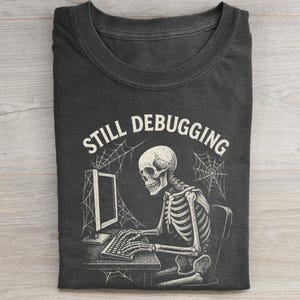 Puede incluir: Camiseta gris oscuro con un esqueleto sentado frente a una computadora, con las palabras "STILL DEBUGGING" arriba. El diseño incluye telarañas y una estética vintage. La camiseta está doblada.