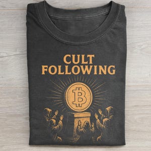 Könnte beinhalten: Dunkelgraues T-Shirt mit dem orangefarbenen Schriftzug "CULT FOLLOWING" über einem Bitcoin-Symbol. Das Bitcoin ist von Lichtstrahlen und Figuren in Kapuzenroben umgeben.
