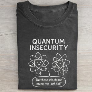 Könnte beinhalten: Dunkelgraues T-Shirt mit dem Text "QUANTUM INSECURITY" und einer Cartoon-Illustration von zwei Atomen. Darunter steht der Text "Do these electrons make me look fat?"