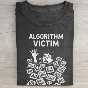 May include: Dark gray t-shirt with the text "ALGORITHM VICTIM" in white. The graphic shows a person drowning in a sea of "AD" and "BUY NOW" signs. The person is reaching out with one hand.