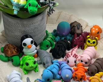 May include: A collection of colorful, 3D-printed animal figurines, including a penguin, frog, seal, and octopus. The figurines are arranged on a burlap surface, with a potted succulent plant in the background. The animals are in various colors.