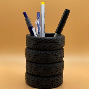 Könnte beinhalten: Ein schwarzer Stiftehalter, der aus einem Stapel von vier Miniatur-Gummireifen besteht. Der zylindrische Behälter enthält vier Schreibgeräte, darunter einen blauen Stift, einen durchsichtigen Kunststoffstift, einen silbernen Marker und einen dicken schwarzen Marker.