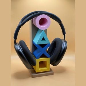 Op de afbeelding: Zwarte over-ear koptelefoon op een kleurrijke geometrische standaard. De standaard heeft een roze cirkel, een turquoise driehoek, een blauwe X en een gele vierkant. De standaard is bruin en de achtergrond is lichtoranje.