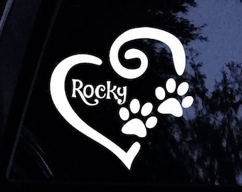 Pode incluir: Adesivo de vinil branco em forma de coração com a palavra "Rocky" dentro e pegadas de patas do lado direito. O adesivo está em uma janela de carro preto.