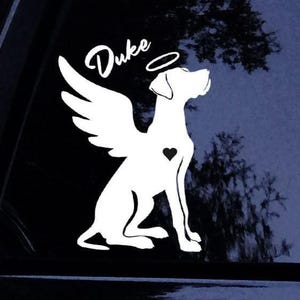 May include: A white vinyl decal featuring a silhouette of a dog with angel wings and a halo above its head. The name Duke is written in a script font above the halo, and a small solid heart is positioned on the chest of the dog figure.