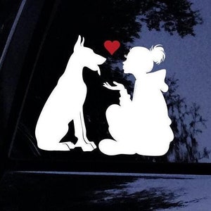 May include: A white vinyl decal featuring the silhouette of a dog sitting opposite a person with a ponytail. The person has an outstretched hand toward the dog, and a small red heart is positioned in the space between their faces.