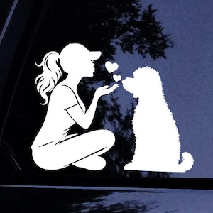 Puede incluir: Un adhesivo de vinilo blanco con la silueta de una persona con gorra de béisbol y coleta, sentada y lanzando besos hacia un perro de pelo rizado. Dos pequeñas formas de corazón flotan entre la persona y el perro sobre un fondo oscuro.