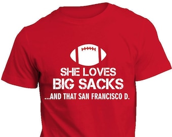 Może przedstawiać: Czerwona koszulka z białym tekstem i grafiką piłki nożnej. Tekst brzmi: "SHE LOVES BIG SACKS...AND THAT SAN FRANCISCO D."