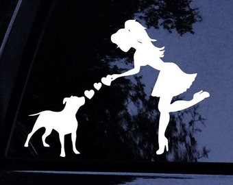 Puede incluir: Pegatina de silueta blanca de una mujer lanzando corazones a un perro. La mujer lleva un vestido y tiene el pelo recogido hacia atrás. El perro es un pitbull. La pegatina está en la ventana de un coche negro.