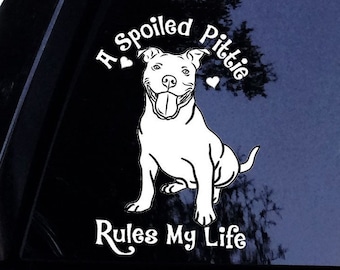 Pode incluir: Um adesivo de vinil branco aplicado em um vidro escuro de carro, com uma ilustração de linhas de um cão pitbull sentado com a língua de fora. O texto A Spoiled Pittie está acima do cão e Rules My Life abaixo, com dois pequenos corações.