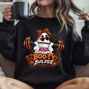 Op de afbeelding: Een zwart sweatshirt met lange mouwen en een afbeelding van een witte geest met een luipaardprint hoofdband en hartvormige zonnebril, die een halter optilt. De tekst luidt BOO-TY BUILDER, omringd door oranje strikken, pompoenen en vleermuizen.