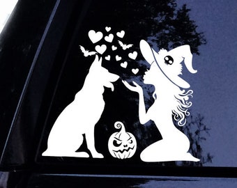 May include: White vinyl decal featuring a witch blowing hearts and bats towards a dog. A jack-o'-lantern sits below the witch. The witch is wearing a hat. The dog is sitting. Halloween themed.