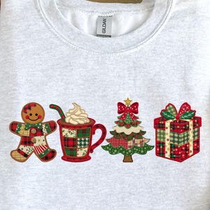 May include: A light grey sweatshirt with festive Christmas designs. It features a gingerbread man, a mug of hot chocolate, a Christmas tree, and a wrapped gift. The designs use red, green, and white plaid patterns, ideal for the holiday season.