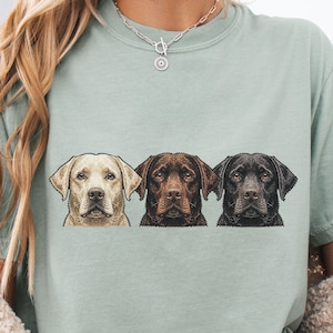 May include: A light green t-shirt featuring a graphic of three Labrador Retrievers: a yellow, a chocolate, and a black lab. The dogs are depicted from the chest up, with detailed fur and facial features.