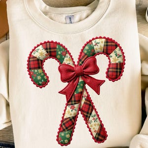 Puede incluir: Sudadera de color crema con un diseño festivo de bastones de caramelo. Los bastones de caramelo están adornados con patrones de cuadros rojos, verdes y dorados y un lazo rojo. Una prenda cómoda y elegante para la temporada navideña.
