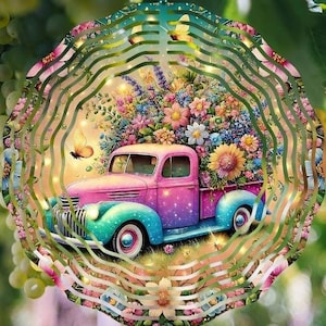 May include: A colorful metal wind spinner with a whimsical design featuring a pink and blue pickup truck carrying a bed of flowers. The truck has a white steering wheel and headlights. The wind spinner has a layered design with multiple circles and swirls.