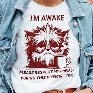 May include: White t-shirt featuring a red cartoon of a cat with messy hair holding a coffee cup. The text on the shirt reads "I'm Awake. Please respect my privacy during this difficult time."