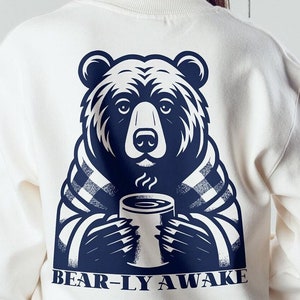May include: White sweatshirt with a navy blue bear design holding a cup of coffee. The text "BEAR-LY AWAKE" is printed below the bear.