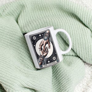 Pu&ograve; includere: Tazza in ceramica bianca con un'illustrazione in bianco e nero di due mani che si tengono. L'illustrazione &egrave; circondata da un cerchio di stelle e il testo "THE LOVERS" &egrave; stampato sotto l'immagine.