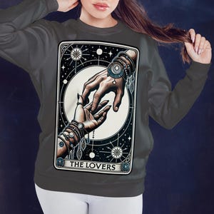 Pu&ograve; includere: Una felpa grigia con una grafica della carta dei Tarocchi degli Amanti. La carta presenta due mani che si raggiungono, circondate da stelle e una luna crescente. Il testo "THE LOVERS" &egrave; stampato nella parte inferiore della carta.