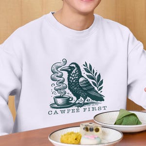 K&ouml;nnte beinhalten: Wei&szlig;es Sweatshirt mit einer schwarzen und gr&uuml;nen Illustration eines Rabens, der Kaffee trinkt. Der Text "CAWFFEE FIRST" ist unter der Illustration gedruckt.