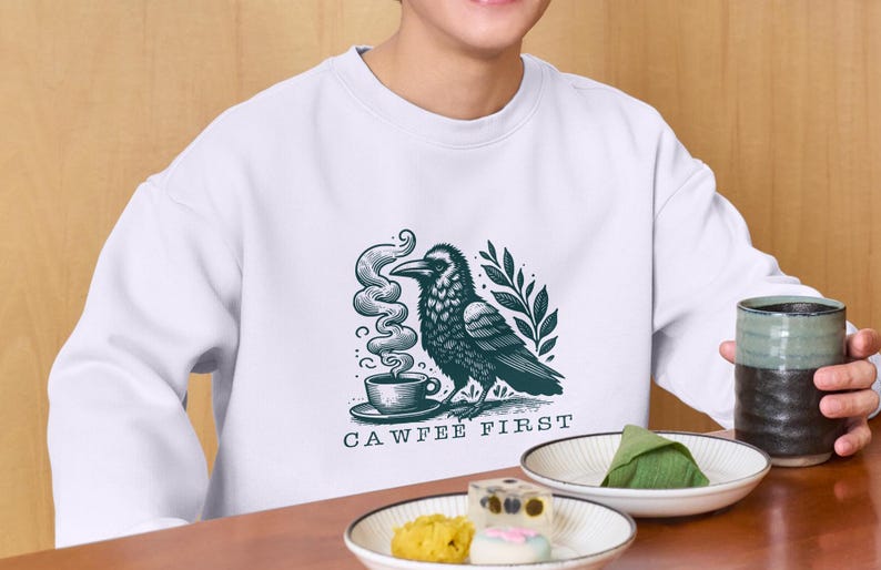 K&ouml;nnte beinhalten: Wei&szlig;es Sweatshirt mit einer schwarzen und gr&uuml;nen Illustration eines Rabens, der Kaffee trinkt. Der Text "CAWFFEE FIRST" ist unter der Illustration gedruckt.