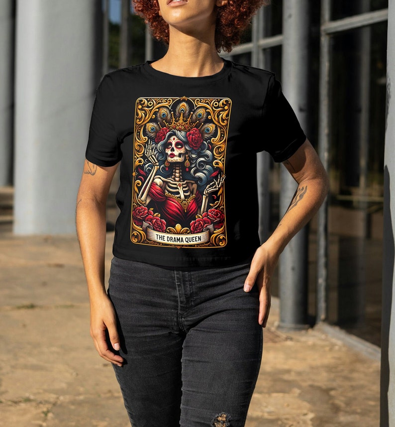 May include: Black t-shirt with a graphic print of a skeleton woman wearing a crown and red dress. The graphic is surrounded by a gold frame and the text "THE DRAMA QUEEN".