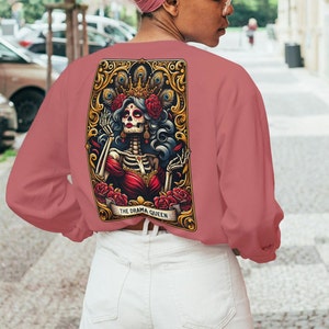 May include: A pink long-sleeved sweatshirt with a graphic print of a skeleton queen wearing a crown and holding a flower. The graphic is framed in gold and has the text "THE DRAMA QUEEN".