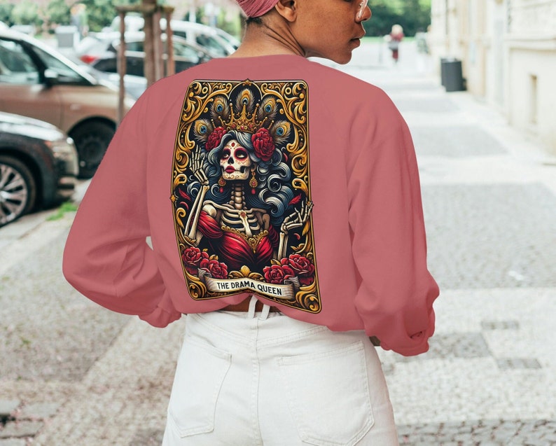 May include: A pink long-sleeved sweatshirt with a graphic print of a skeleton queen wearing a crown and holding a flower. The graphic is framed in gold and has the text "THE DRAMA QUEEN".