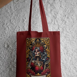 May include: A maroon tote bag with a colorful illustration of a skeleton woman wearing a crown and a red dress. The illustration is surrounded by a gold frame with peacock feathers and roses. The text "THE DRAMA QUEEN" is printed below the illustration.