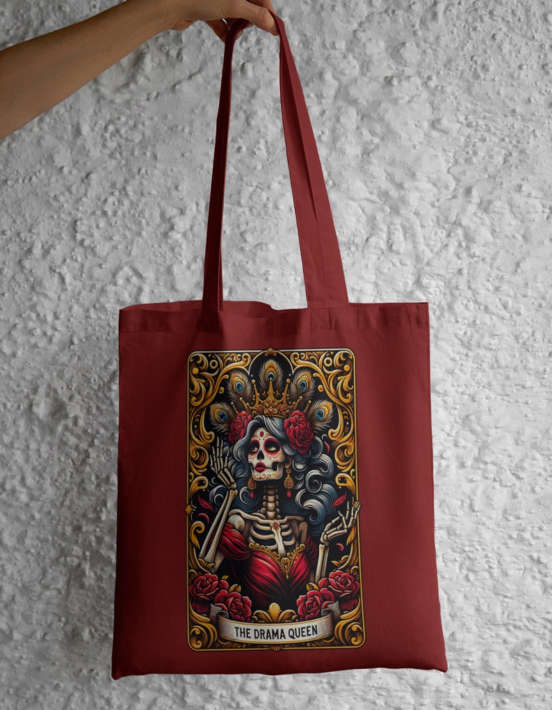 May include: A maroon tote bag with a colorful illustration of a skeleton woman wearing a crown and a red dress. The illustration is surrounded by a gold frame with peacock feathers and roses. The text "THE DRAMA QUEEN" is printed below the illustration.