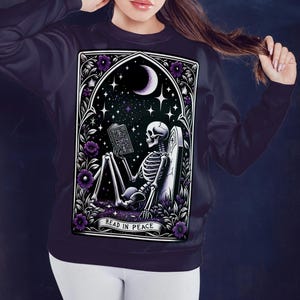 May include: A dark purple sweatshirt with a graphic print of a skeleton reading a book in a graveyard setting. The graphic is framed in a gothic archway with purple flowers and the text "Read in Peace".