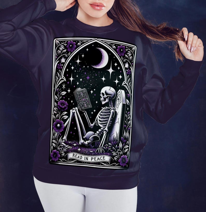 May include: A dark purple sweatshirt with a graphic print of a skeleton reading a book in a graveyard setting. The graphic is framed in a gothic archway with purple flowers and the text "Read in Peace".