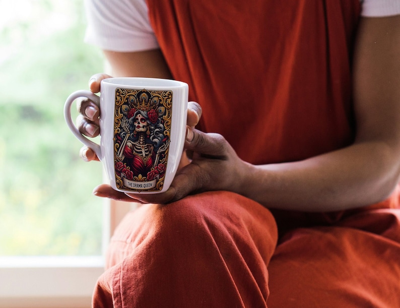 May include: White ceramic mug with a colorful illustration of a skeleton queen with a crown and a red dress. The mug has the text "The Drunk Queen" printed on the bottom.