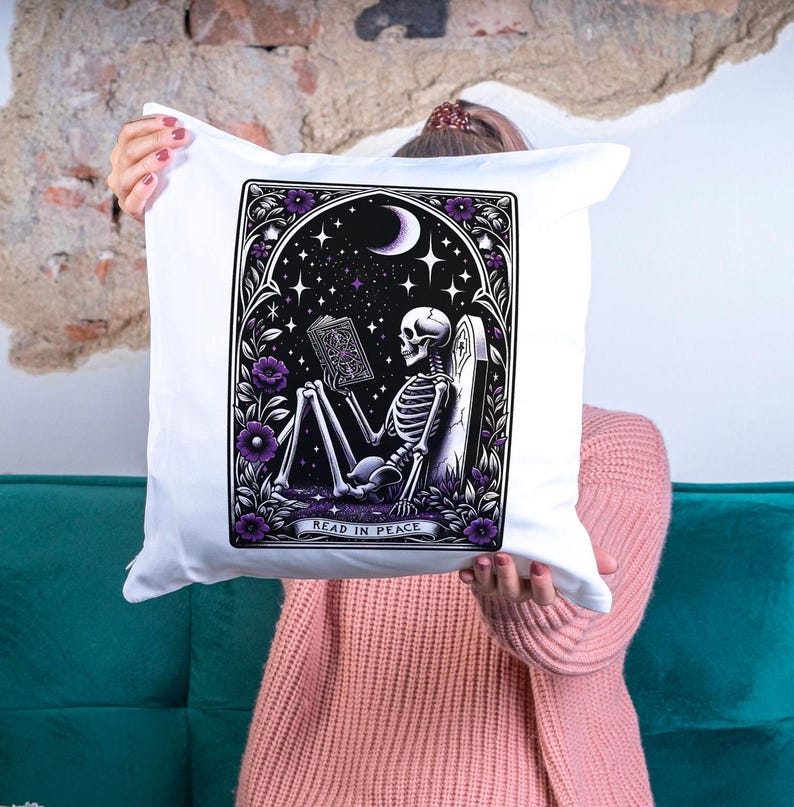 May include: White pillow with a black and white illustration of a skeleton reading a book in a graveyard. The illustration is surrounded by a purple floral border. The text "Read in Peace" is below the illustration.