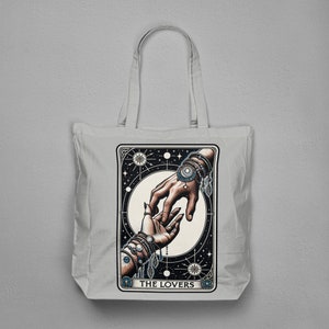 Pu&ograve; includere: Una borsa tote in tela bianca con un'illustrazione in bianco e nero della carta dei Tarocchi degli Amanti. La carta mostra due mani che si raggiungono, adornate con gioielli. Il testo "THE LOVERS" &egrave; stampato nella parte inferiore della carta.
