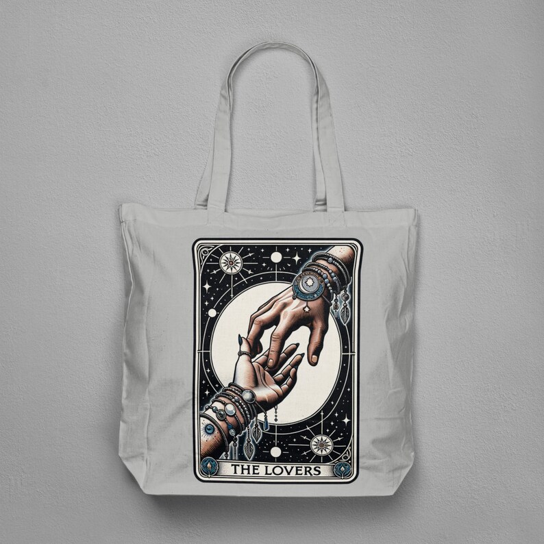 Pu&ograve; includere: Una borsa tote in tela bianca con un'illustrazione in bianco e nero della carta dei Tarocchi degli Amanti. La carta mostra due mani che si raggiungono, adornate con gioielli. Il testo "THE LOVERS" &egrave; stampato nella parte inferiore della carta.