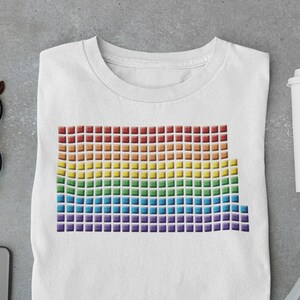 May include: A white t-shirt featuring a pixelated rainbow design. The design is made up of small squares in shades of red, orange, yellow, green, blue, indigo, and violet.