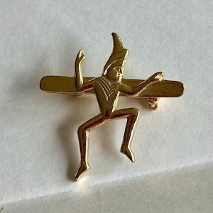 May include: A gold-colored metal pin featuring a stylized figure with outstretched arms and a pointed hat, mounted on a horizontal bar. The pin has a classic, vintage aesthetic.