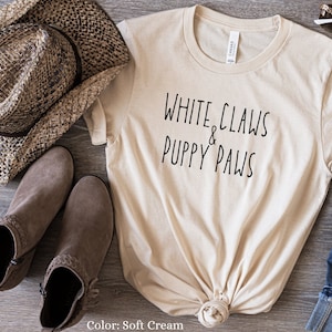 Könnte beinhalten: Ein T-Shirt in zartem Cremeton mit dem Aufdruck "White Claws & Puppy Paws" in Schwarz. Das Shirt ist unten verknotet und liegt auf einer Holzoberfläche mit einem braunen Cowboyhut, braunen Stiefeln und einer blauen Jeans.
