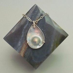 Peut inclure: Un collier pendentif en forme de goutte d'eau avec une grande perle irisée sertie dans un cadre argenté. Le pendentif est suspendu à une chaîne en argent. Le collier est présenté sur une surface sombre et géométrique.