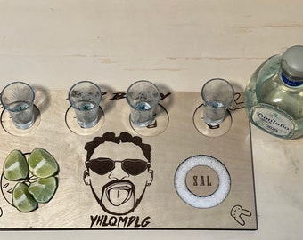 May include: Wooden shot glass tray with four shot glasses, a bottle of tequila, lime wedges, and a salt dispenser. The tray features a cartoon image of a man with the text "YHLQMDLG" underneath.