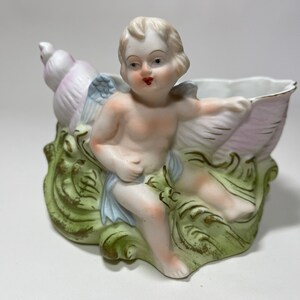May include: A porcelain cherub figurine seated in a shell-shaped planter. The cherub has blonde hair, rosy cheeks, and red lips. The shell is white and pink, with green and gold accents. The planter is a decorative item.