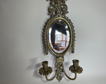 May include: An ornate brass wall mirror with two candle holders. The mirror is oval shaped and has a decorative frame. The candle holders are on either side of the mirror and have a floral design.
